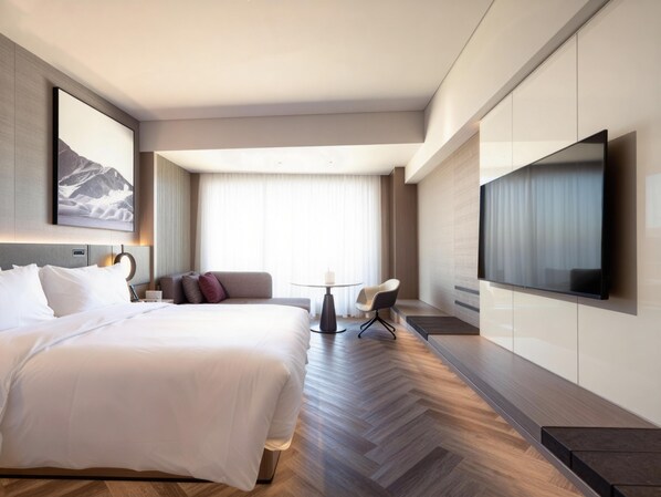 A modern hotel room with a large bed, a flat-screen TV, a sofa, a small table, and a large window with curtains.