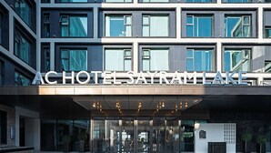 Una entrada de hotel moderna con fachada de vidrio transparente, un letrero que dice 'ACHOTEL SAYRAMLAKE' y un área de acceso pavimentada.