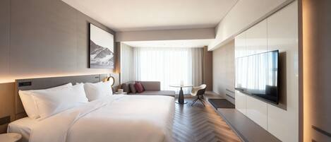 A modern hotel room with a large bed, a TV, a sofa, and a dining table.