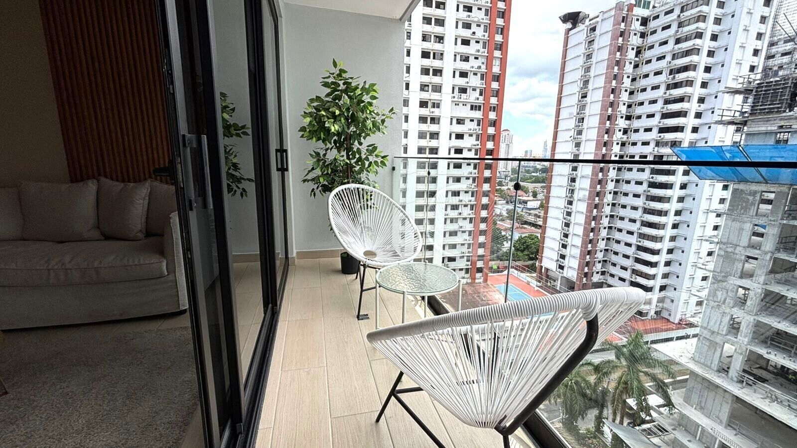 Uma varanda com sofá, duas cadeiras, uma mesinha e vista para edifícios altos.