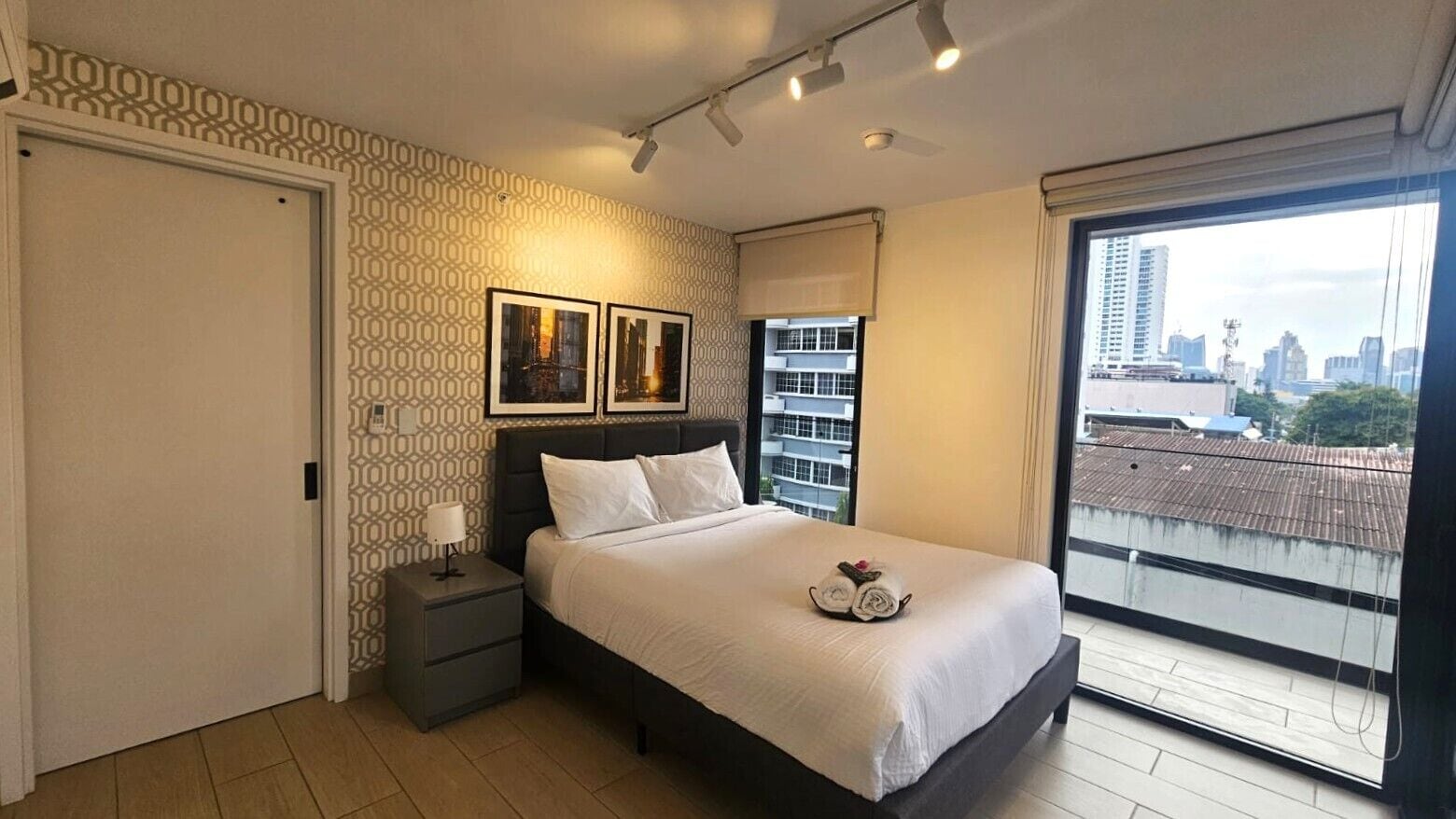 Een slaapkamer met een bed, een nachtkastje, een raam met uitzicht op de stad en behang met een patroon.