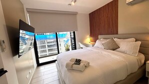 A hotel room with a bed, a TV, a window with a view of buildings, and a wooden panel on the wall. - Living 73 Panamá by Boutique Apartments (Panama City)
