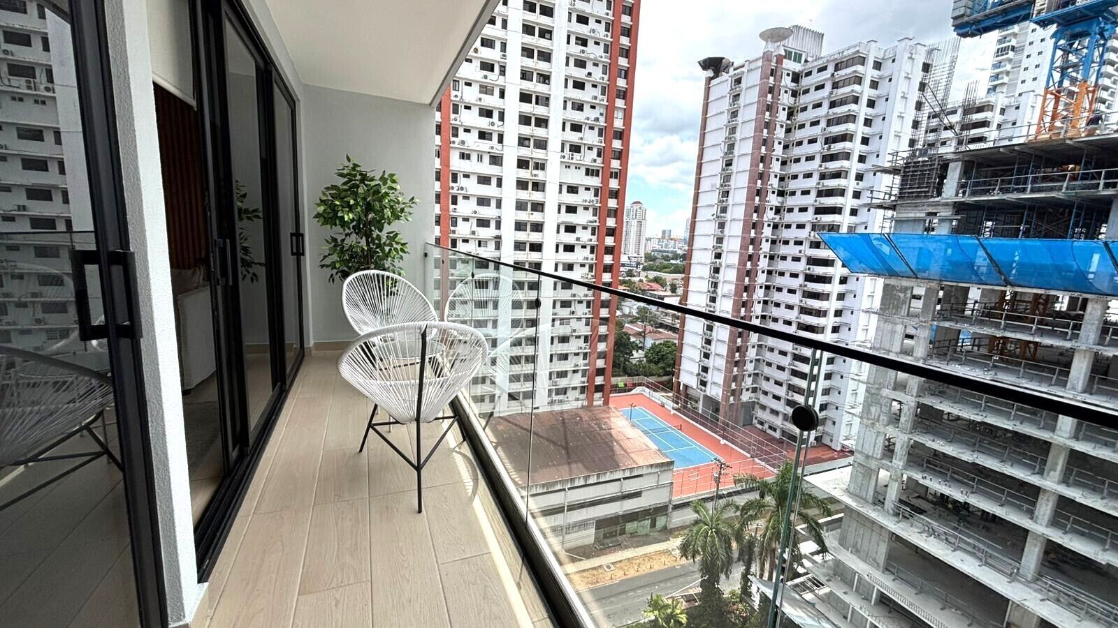 Un balcón con una silla, una planta en maceta y vistas de edificios altos y una obra en construcción.