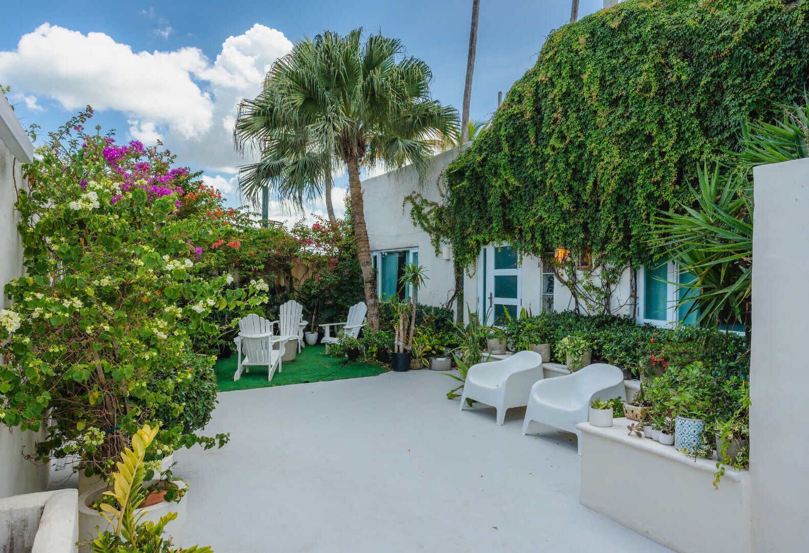 A patio with white chairs and tables, surrounded by lush greenery and flowering plants.