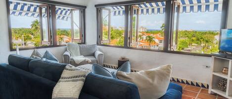 Una sala de estar con un sofá azul oscuro, un sillón blanco y un televisor sobre un mueble bajo. Amplias ventanas con toldos a rayas ofrecen vistas a un pueblo costero y palmeras.