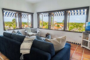 Una sala de estar con un sofá azul oscuro, un sillón blanco y un televisor sobre un mueble bajo. Amplias ventanas con toldos a rayas ofrecen vistas a un pueblo costero y palmeras.