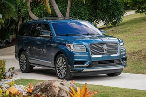Un SUV azul estacionado en una entrada de casa con un camino de piedra y plantas tropicales al fondo.