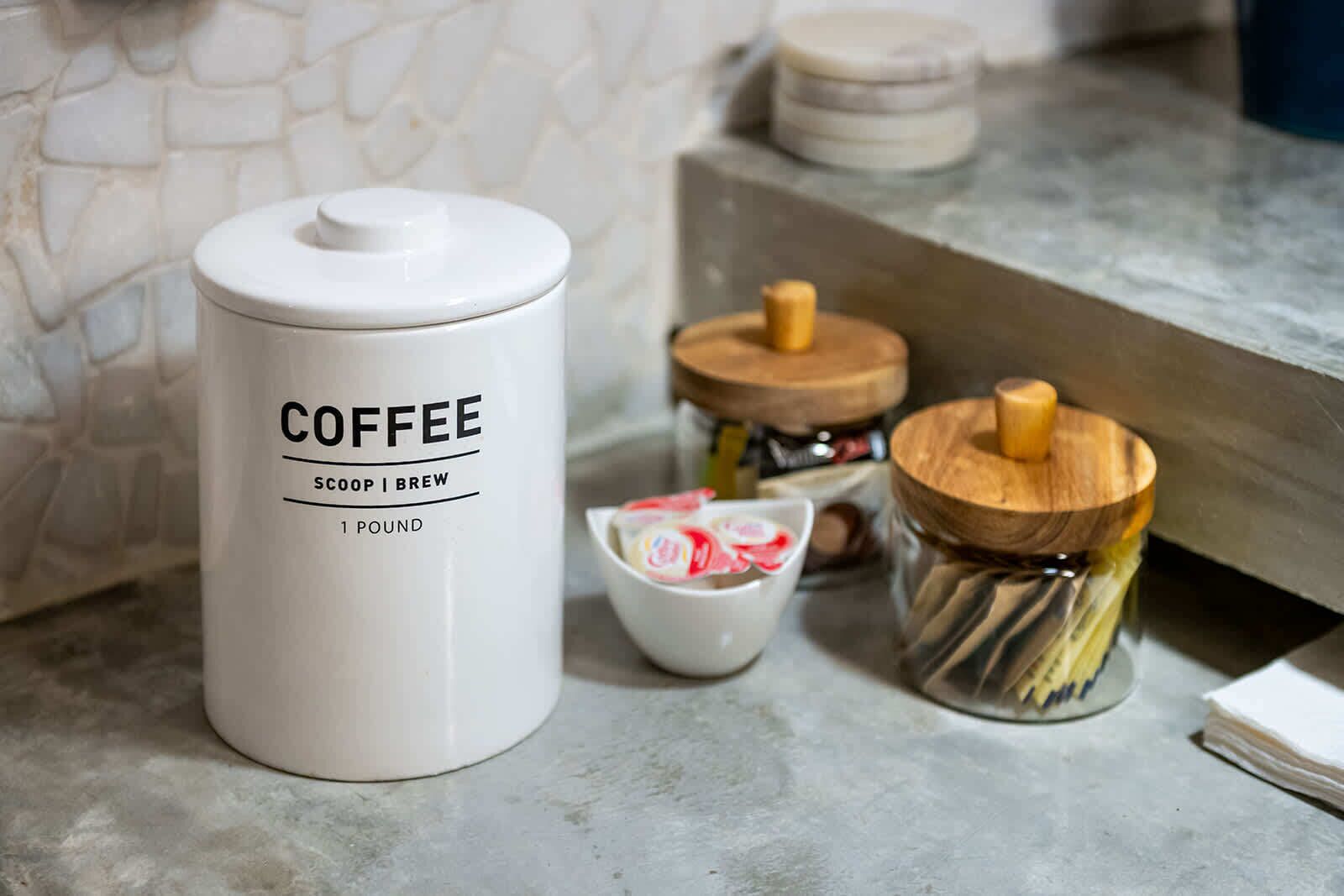 Un plan de travail de cuisine avec un récipient à café étiqueté « COFFEE SCOOP BREW 1 POUND » (café moulu, 1 livre), un petit bol et deux pots en bois avec des couvercles.