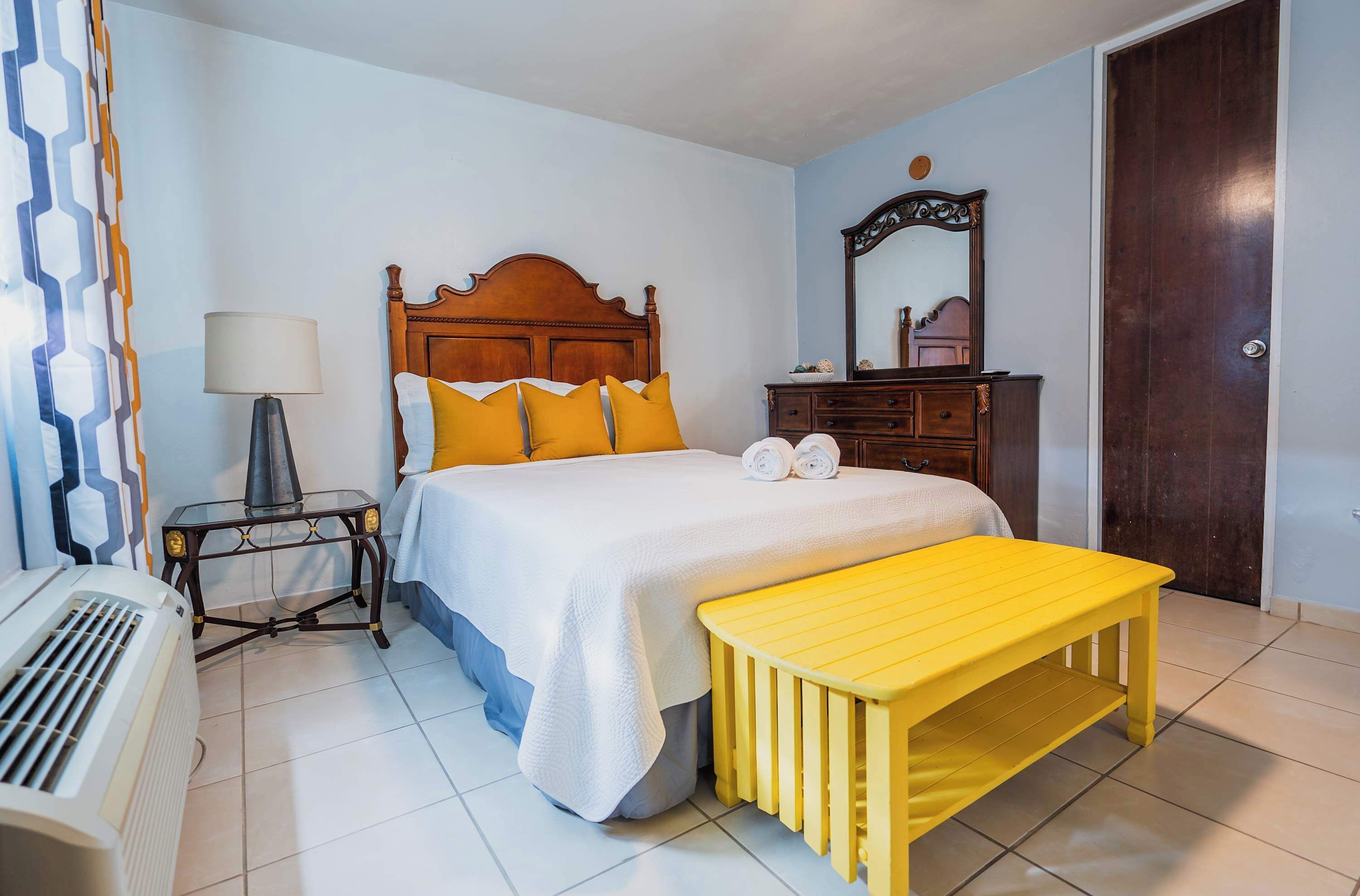 A bedroom with a wooden bed, a bedside table with a lamp, a bench, and a dresser.