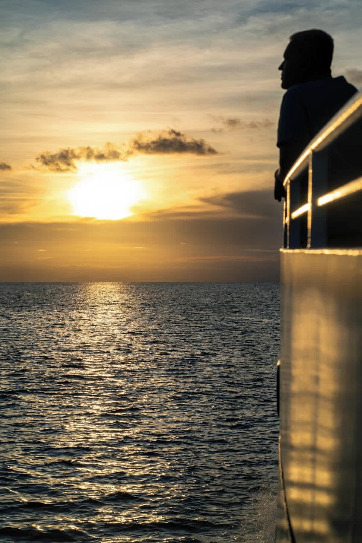 Una persona parada en un balcón, con vista a la puesta de sol sobre el océano.