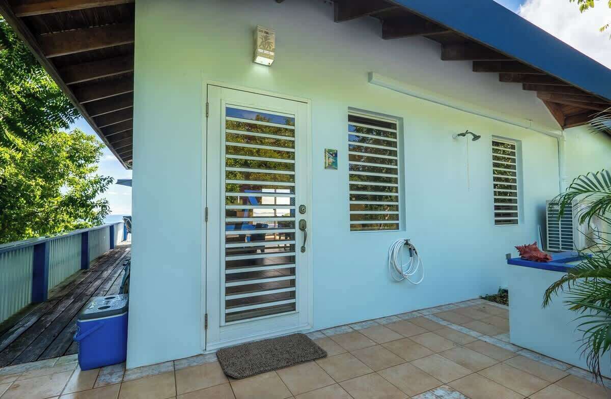 A covered outdoor area with a tiled floor, a door with a metal grill, and a small blue trash bin.