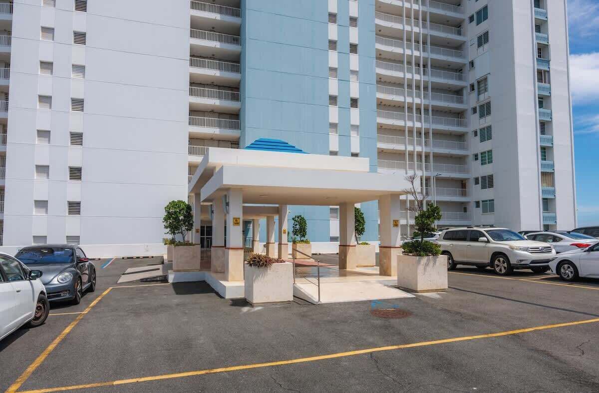 A modern hotel entrance with a covered waiting area, multiple floors, and a parking lot with several cars.