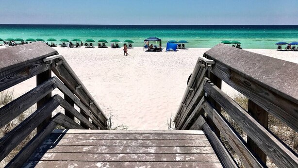 Eine Holztreppe führt zu einem Strand mit grünen Sonnenschirmen und Liegestühlen.