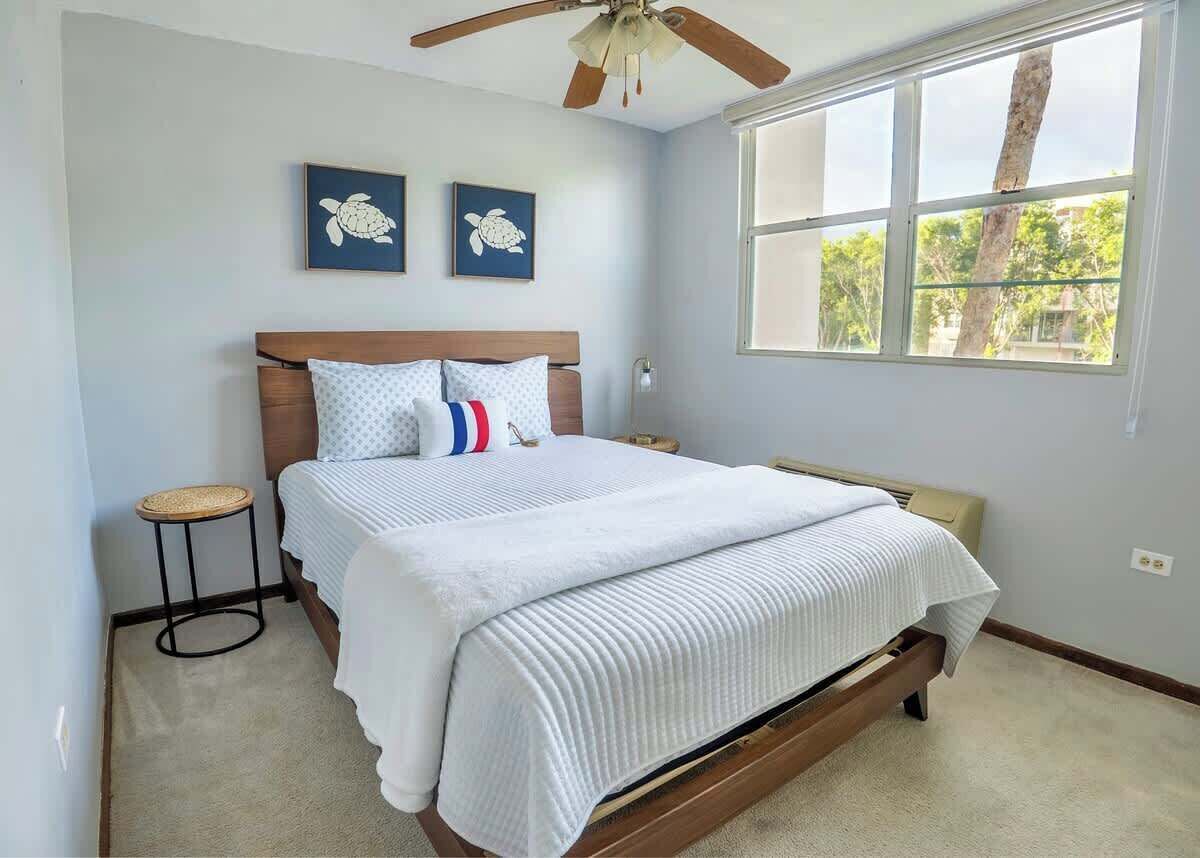A bedroom with a bed, a ceiling fan, two wall art pieces, a window, and a small stool.