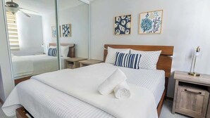 A neatly made bed with white bedding and a rolled towel, a wooden nightstand with a lamp, and two wall art pieces depicting leaves. - Serenity Beach Vibe Stay in Costa Brava, Ceiba (Ceiba)