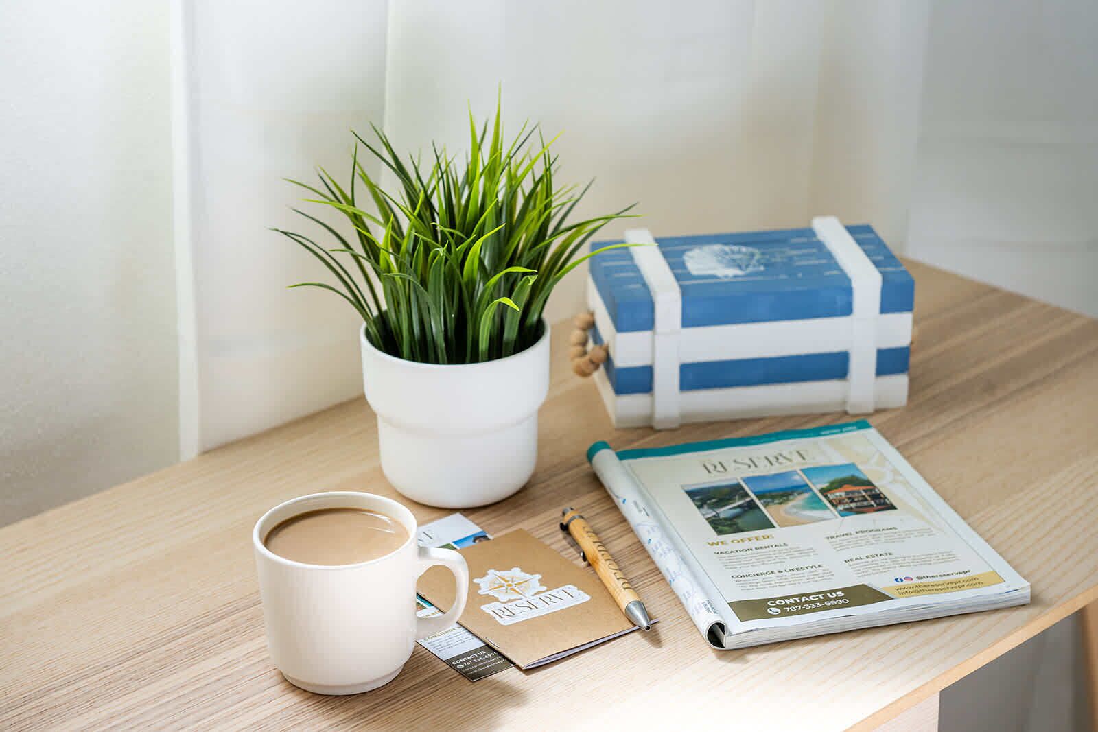 A wooden table with a white mug, a small potted plant, a brochure, a pen, and a small card.