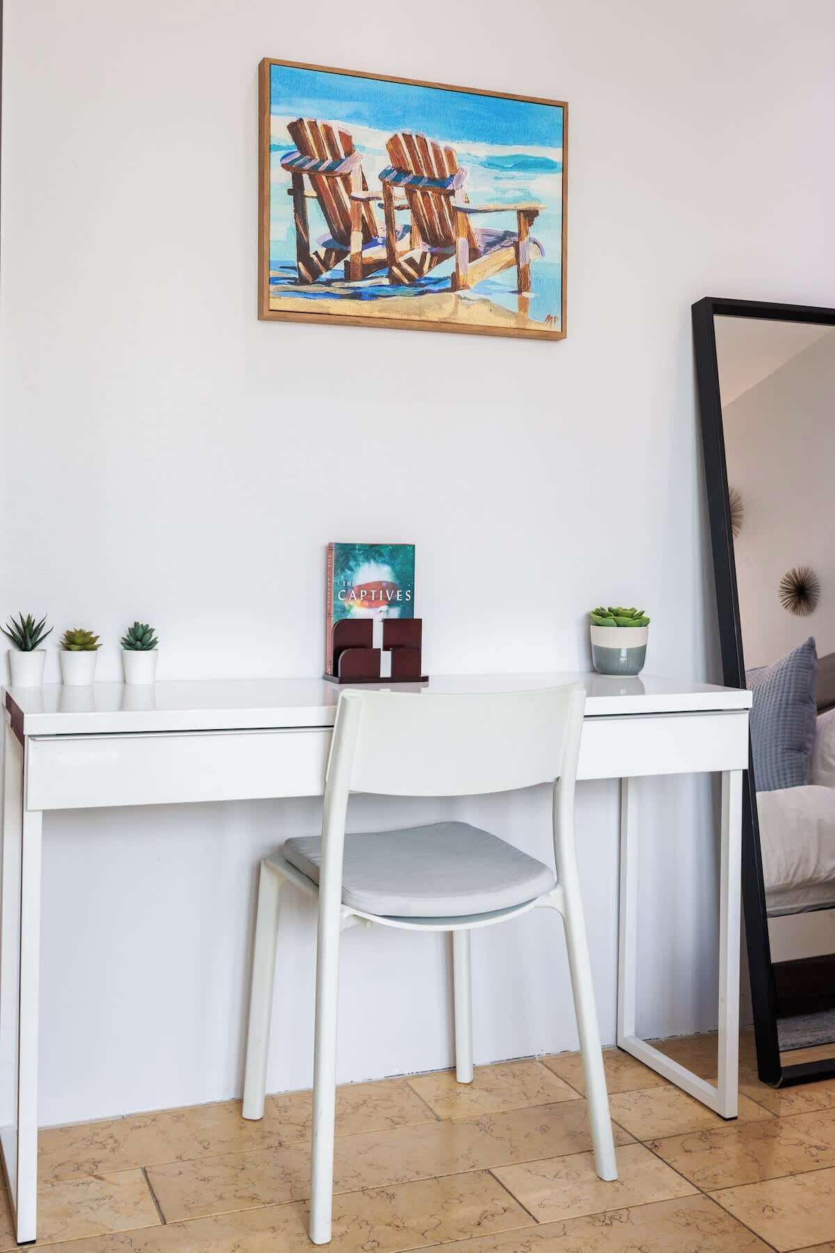 A white desk with a chair, a small potted plant, a book titled 'The Captives', and a painting of chairs on a beach.