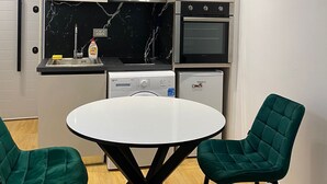 Ett modernt kök med vita skåp, en svart marmorvägg bakom arbetsbänken, en inbyggd ugn och en diskmaskin. Där finns ett runt bord med två gröna stolar.
