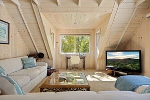 A cozy attic room with a wooden ceiling, a large window, a TV, a sofa, and a coffee table. - Home Sweet Home, Lakefront Vibes w/Dock & Hot Tub (Big Bear Lake)