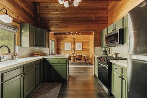 A kitchen with wooden walls, green cabinets, a central island, and a dining area with chairs. - Shadow Wood Lodge, w Hot Tub near Ski Slopes (Big Bear Lake)