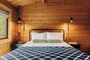 A wooden cabin bedroom with a metal bed frame, two bedside lamps, a patterned bedspread, and a window with a view. - Shadow Wood Lodge, w Hot Tub near Ski Slopes (Big Bear Lake)