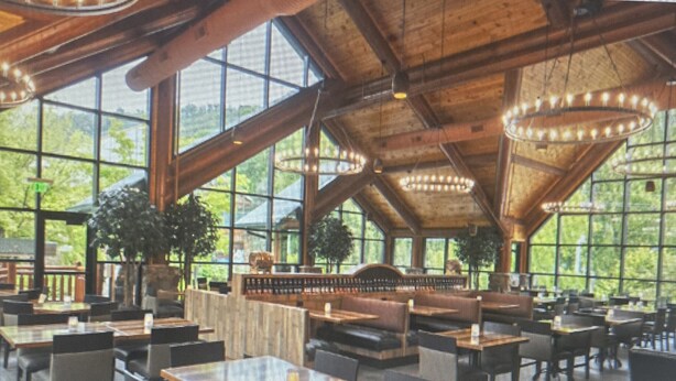 Ein geräumiges Restaurant mit hohen Decken, großen Fenstern und Holzbalken. Tische und Stühle sind für das Essen angerichtet.