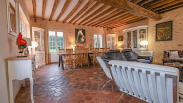 Un intérieur rustique avec des poutres apparentes, un sol en pierre et un coin repas comprenant des chaises en bois et une table blanche.