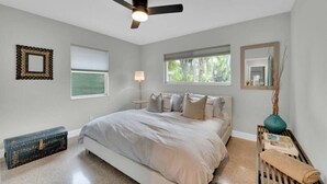 Quarto com cama, ventoinha no teto, janela, mesa de cabeceira com abajur e espelho decorativo.