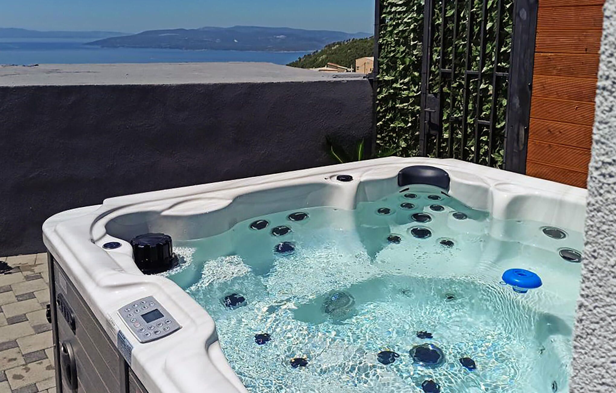 A hot tub with a control panel, surrounded by a wooden fence and a view of the sea.