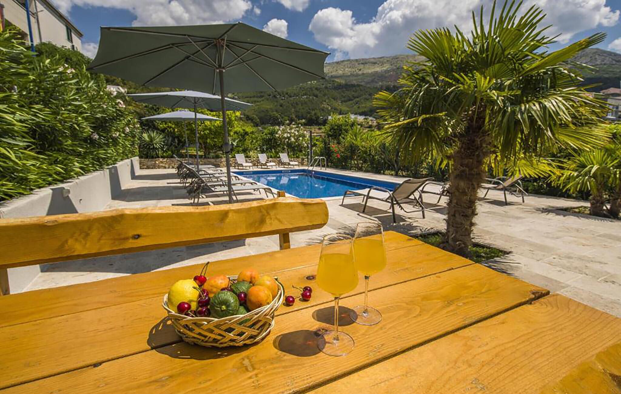 Un tavolo di legno con un cesto di frutta e due bicchieri di una bevanda gialla, una piscina e sedie a sdraio in un ambiente esterno soleggiato.