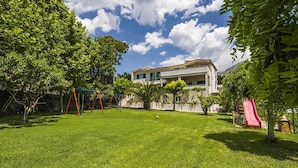 A well-maintained grassy backyard with a swing set, a small palm tree, and a building with a balcony. - Cozy home in Podstrana with private swimming pool, can be inside or outside (Podstrana)