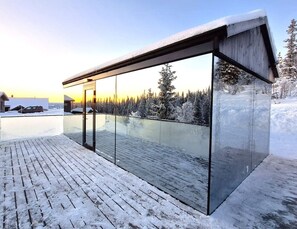Een moderne glazen constructie met een houten terras, gelegen tegenover een besneeuwd landschap met bomen en een zonsondergang.