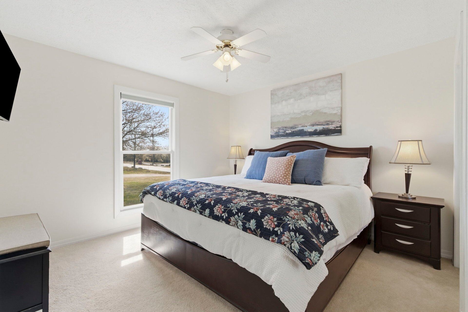 A bedroom with a large bed, a ceiling fan, a nightstand with a lamp, and a window with a view of trees.