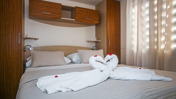 A neatly made bed with two white towels arranged in a heart shape, a wooden headboard, and a window with sheer curtains. - Gorgeous (sta)caravan in Drnis with WiFi (Drnis)
