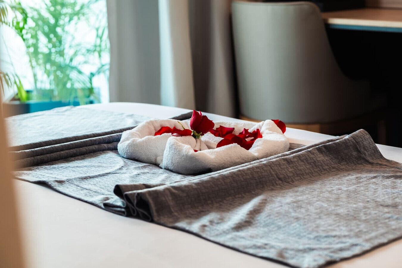 Un lit bien fait, recouvert d’une couverture grise et agrémenté d’un oreiller blanc, parsemé de pétales de roses rouges et surmonté d’une seule rose rouge.