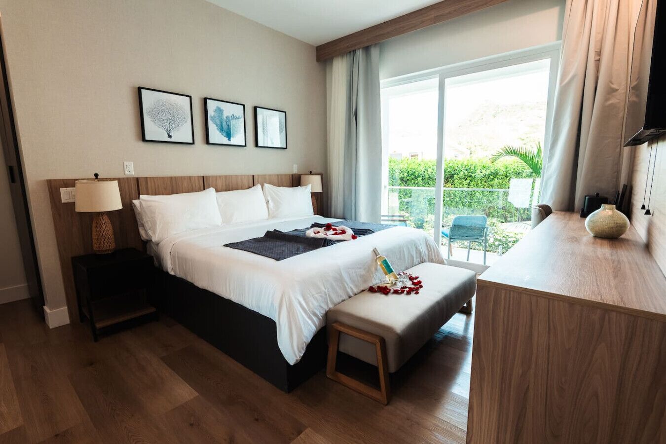 Une chambre d’hôtel avec un grand lit, une table de chevet avec une lampe, un petit banc parsemé de pétales de rose et une vue sur la verdure par la fenêtre.