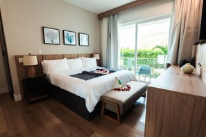 A hotel room with a large bed, a bedside table with a lamp, a small bench with rose petals, and a view of greenery through the window. - 3 King Bed Suite @CockleshellBeach w/View of Nevis (Southeast Peninsula)