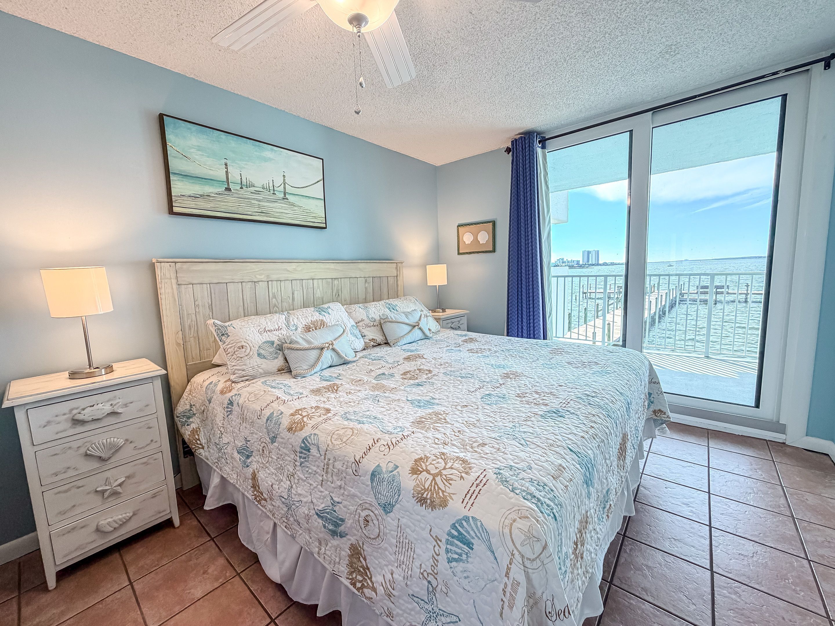 Un dormitorio con una cama grande, una mesita de noche con lámpara, un aparador y vistas al mar desde un balcón.
