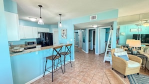 A spacious kitchen with white cabinets, a black refrigerator, and a microwave. There are two bar stools and a dining area with a wicker sofa and a table. - Best Sunsets on the Lagoon Front w/ Pools & Pier (Gulf Shores)