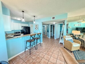 A spacious kitchen with white cabinets, a black refrigerator, and a microwave. There are two bar stools and a dining area with a wicker sofa and a table. - Best Sunsets on the Lagoon Front w/ Pools & Pier (Gulf Shores)