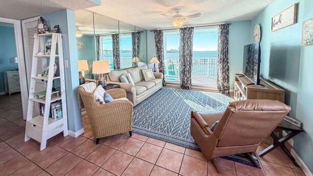 A living room with a sofa, chairs, a TV, a bookshelf, and a large window with a view of the sea. - Best Sunsets on the Lagoon Front w/ Pools & Pier (Gulf Shores)