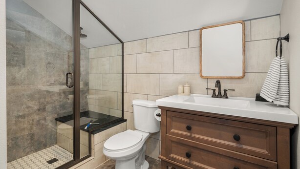 A bathroom with a glass shower enclosure, a white toilet, a wooden vanity with a mirror, and beige tiles. - The South Hill Nook, 4 min to Manito Park (Spokane)