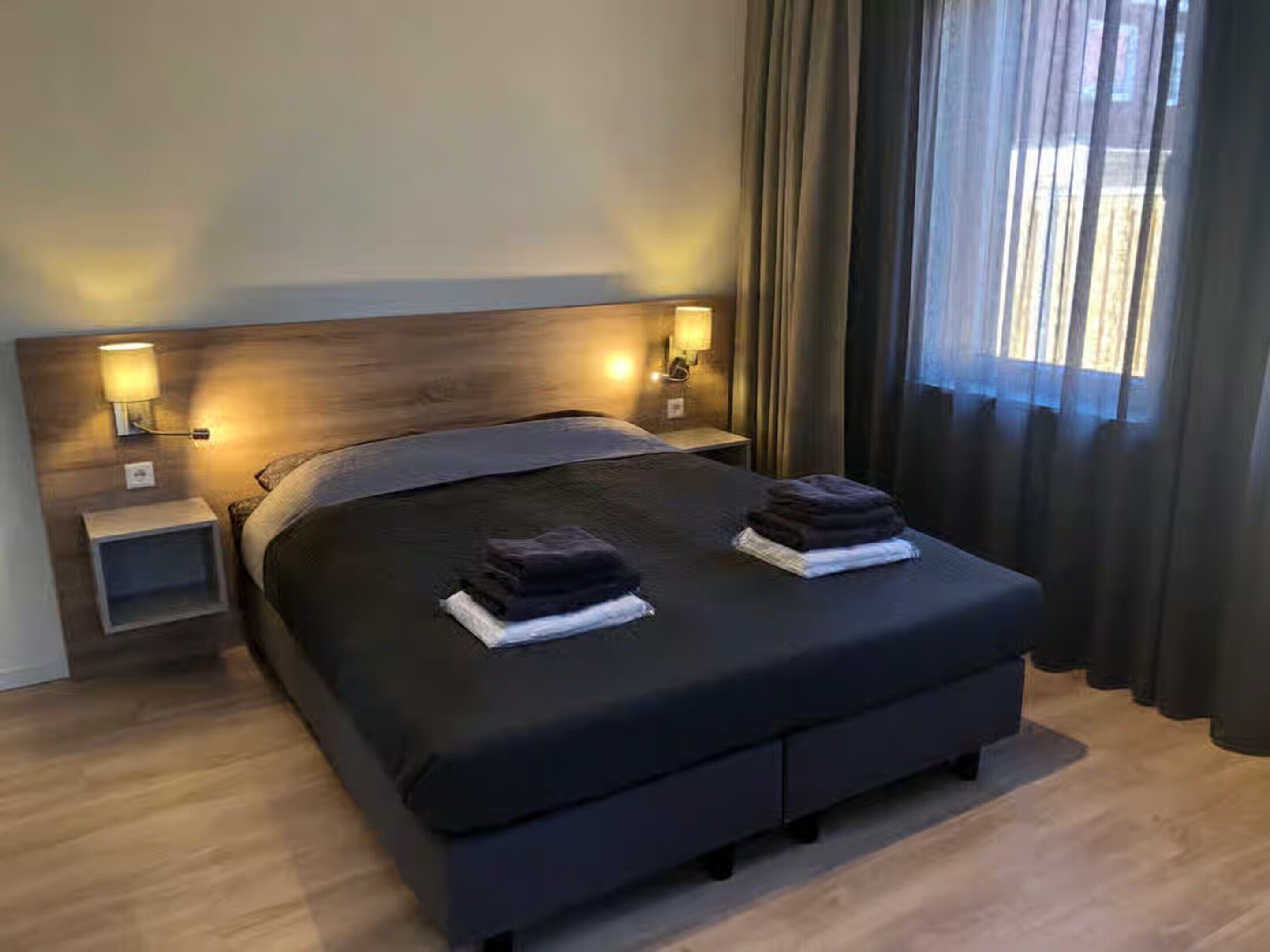 A modern hotel room with a large bed, two bedside tables, and a view of the outdoors through sheer curtains.