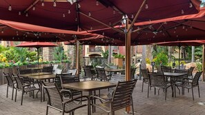 Een terras buiten met tafels en stoelen onder rode parasols, omgeven door groen en een stenen muur.