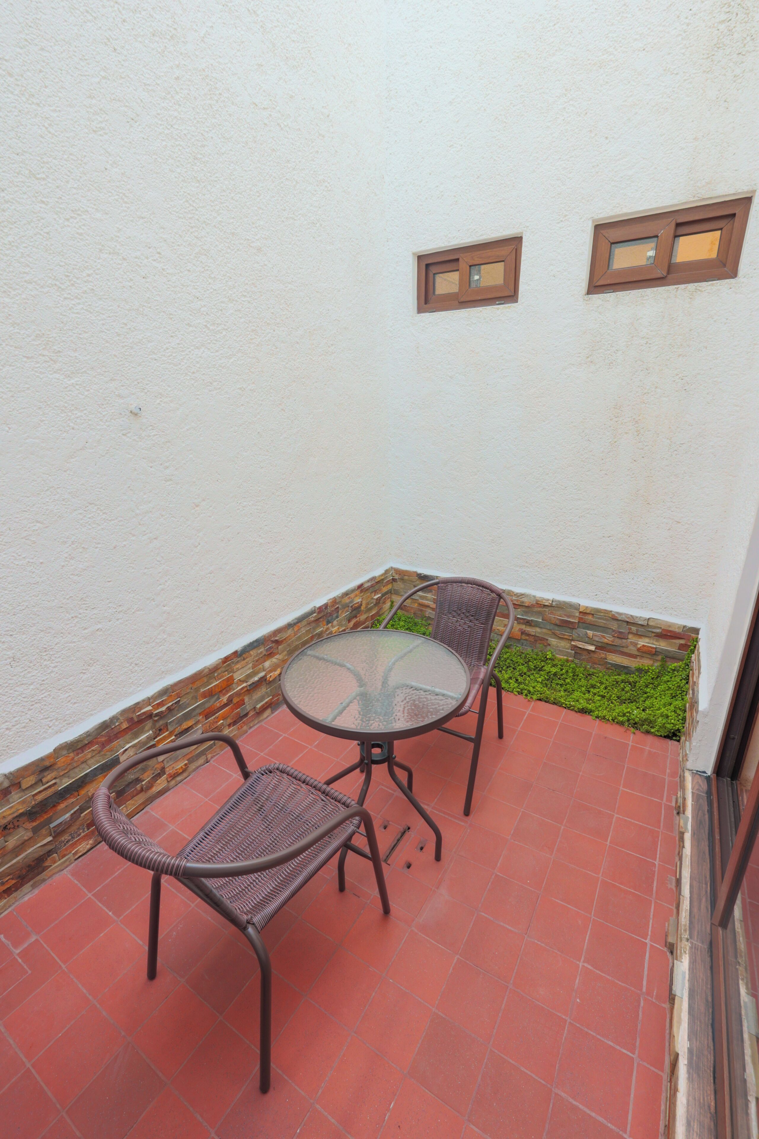A small patio with a round glass table and two wicker chairs, a tiled floor, and a white wall with two small windows.