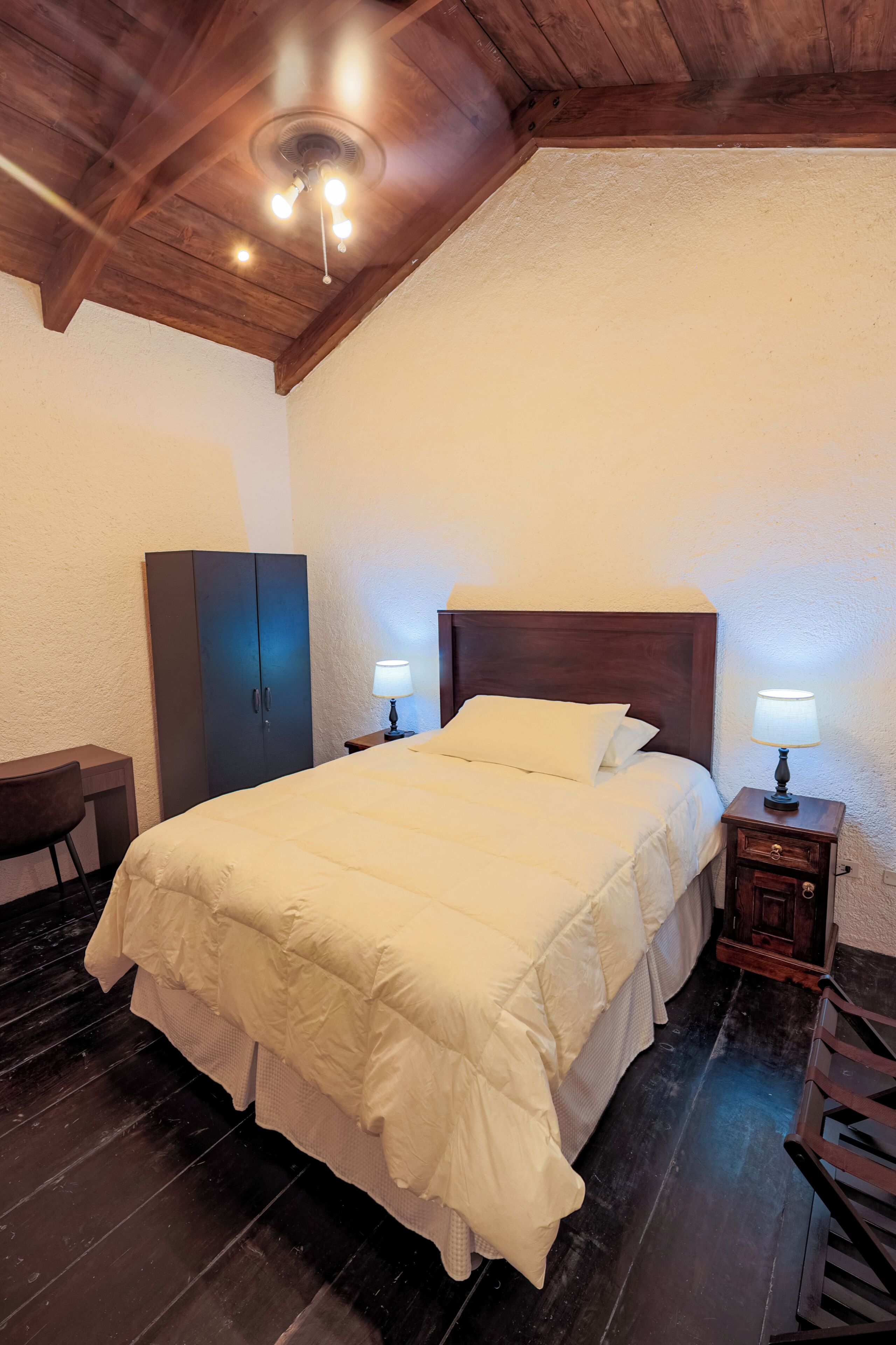 Un dormitorio con techo de madera, cama con ropa de cama blanca, escritorio con silla y dos lámparas de noche.
