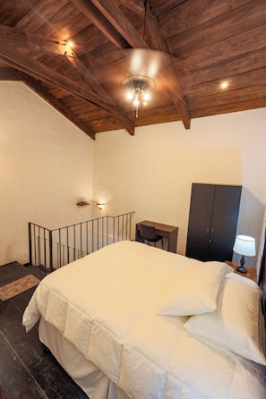 A bedroom with a large bed, wooden ceiling beams, a desk, a wardrobe, and a nightstand with a lamp.