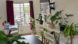 Una habitación con una mesa negra, estanterías blancas con plantas en macetas, una ventana con cortinas y un televisor montado.
