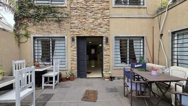 Un patio al aire libre con un muro de piedra, dos ventanas con rejas de metal y una puerta que da al interior. Hay sillas y mesas blancas, y macetas con plantas alrededor.