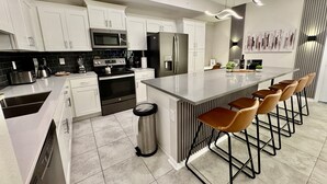 Una cucina moderna con mobili bianchi, elettrodomestici in acciaio inossidabile, un'isola centrale con sgabelli da bar e pavimento piastrellato.